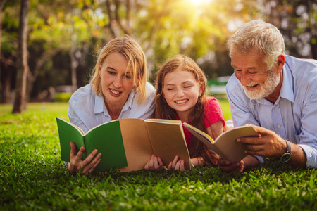 Happy family read books together and lying on green grass in public park. Little girl kid learning with mother and father in outdoors garden. Education and family lifestyle.の写真素材