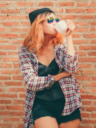 Happy young hipster woman standing against brick wall in town street with coffee cup in her hand.の写真素材