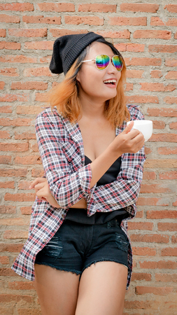 Happy young hipster woman standing against brick wall in town street with coffee cup in her hand.の写真素材
