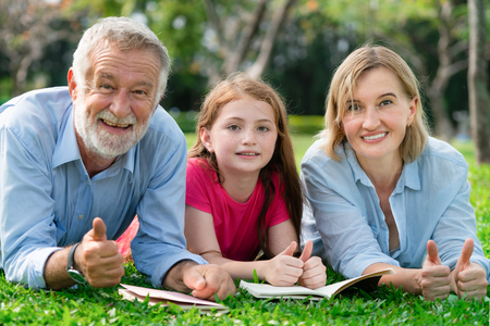 Happy family read books together and lying on green grass in public park. Little girl kid learning with mother and father in outdoors garden. Education and family lifestyle.の写真素材