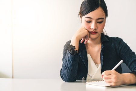 Beautiful young Asian woman writes letter on notebook while sitting at office desk. Content writer...
