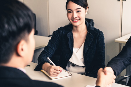 Business people agreement concept. Asian Businessman do handshake with another businessman in the office meeting room. Young Asian secretary lady sits beside him.の写真素材