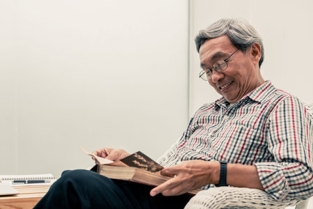 Happy senior Asian man reading book on the chair in living room at home. Retirement lifestyle and...