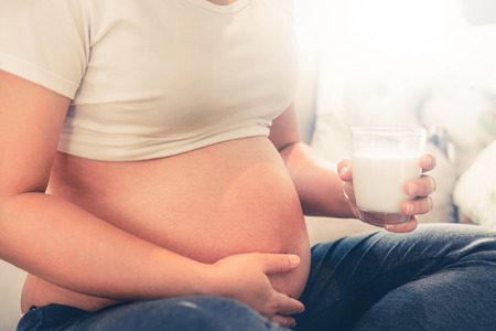 Happy pregnant woman drinks milk in glass at home while taking care of her child. The young expecting mother holding baby in pregnant belly. Calcium food nutrition for strong bones of pregnancy.の写真素材
