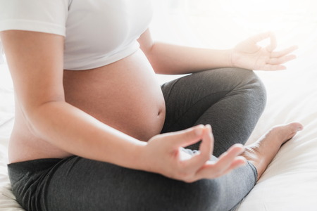 Pregnant woman doing yoga exercise on bed in bedroom at home while taking care of her child. The happy young expecting mother holding baby in pregnant belly. Maternity prenatal care and pregnancy.の写真素材