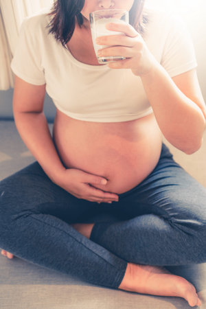 Happy pregnant woman drinks milk in glass at home while taking care of her child. The young...
