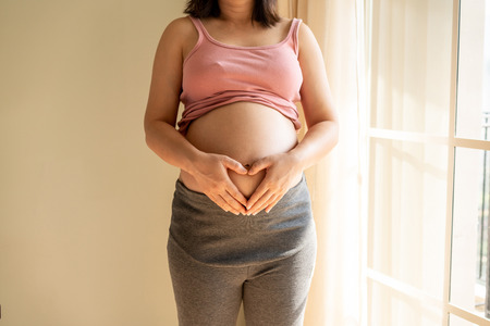 Pregnant woman feeling happy at home while taking care of her child. The young expecting mother holding baby in pregnant belly. Maternity prenatal care and woman pregnancy concept.の写真素材