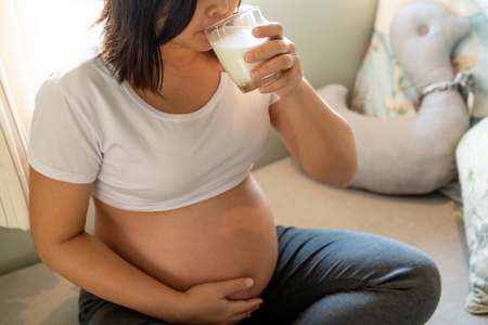 Happy pregnant woman drinks milk in glass at home while taking care of her child. The young expecting mother holding baby in pregnant belly. Calcium food nutrition for strong bones of pregnancy.の写真素材
