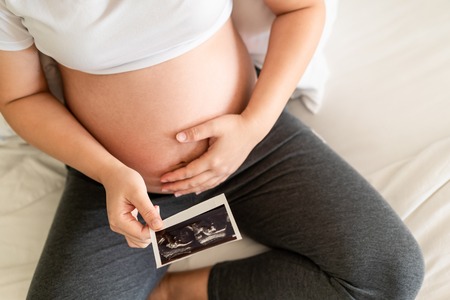 Pregnant woman feeling happy at home while taking care of her child. The young expecting mother holding baby in pregnant belly. Maternity prenatal care and woman pregnancy concept.の写真素材
