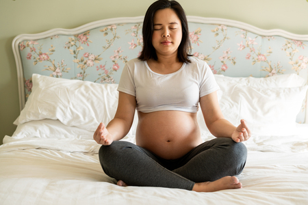 Pregnant woman doing yoga exercise on bed in bedroom at home while taking care of her child. The happy young expecting mother holding baby in pregnant belly. Maternity prenatal care and pregnancy.の写真素材
