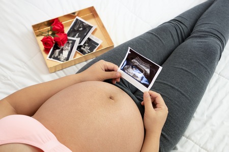 Pregnant woman feeling happy at home while taking care of her child. The young expecting mother holding baby in pregnant belly. Maternity prenatal care and woman pregnancy concept.の写真素材