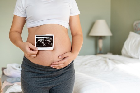 Pregnant woman feeling happy at home while taking care of her child. The young expecting mother holding baby in pregnant belly. Maternity prenatal care and woman pregnancy concept.の写真素材