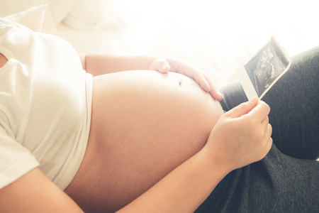 Pregnant woman feeling happy at home while taking care of her child. The young expecting mother holding baby in pregnant belly. Maternity prenatal care and woman pregnancy concept.の写真素材