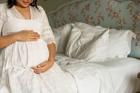 Pregnant woman feeling happy at home while taking care of her child. The young expecting mother holding baby in pregnant belly. Maternity prenatal care and woman pregnancy concept.の写真素材