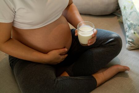 Happy pregnant woman drinks milk in glass at home while taking care of her child. The young expecting mother holding baby in pregnant belly. Calcium food nutrition for strong bones of pregnancy.の写真素材