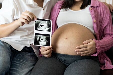 Pregnant couple of husband and wife feels love and relax at home. Young expecting woman holds baby in pregnant belly. Father take care of pregnant mother. Concept of maternity and pregnancy care.の写真素材