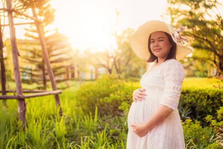 Pregnant woman feeling happy with new life at garden home while take care of her child. The young expecting mother holding baby in pregnant belly. Maternity prenatal care and woman pregnancy concept.の写真素材