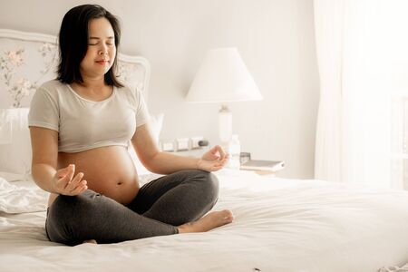 Pregnant woman doing yoga exercise on bed in bedroom at home while taking care of her child. The happy young expecting mother holding baby in pregnant belly. Maternity prenatal care and pregnancy.の写真素材