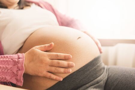 Pregnant woman feeling happy at home while taking care of her child. The young expecting mother holding baby in pregnant belly. Maternity prenatal care and woman pregnancy concept.の写真素材