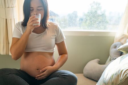 Happy pregnant woman drinks milk in glass at home while taking care of her child. The young expecting mother holding baby in pregnant belly. Calcium food nutrition for strong bones of pregnancy.の写真素材