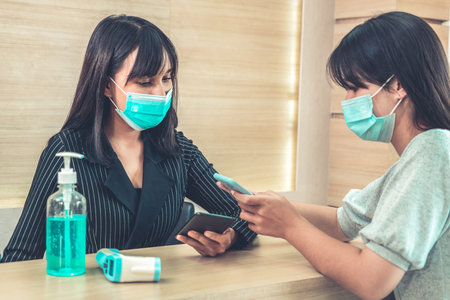 Receptionist and guest wearing face mask at front desk while having conversation in office or...