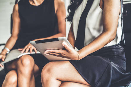 Two businesswomen discussing business while looking at financial data in their hands. They are sitting on the chairs in the office meeting room.の写真素材