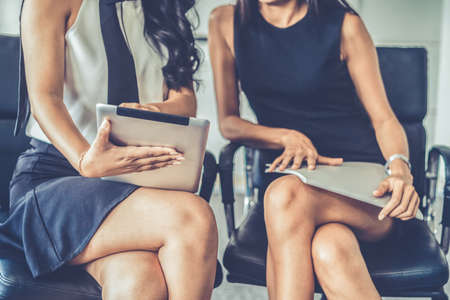 Two businesswomen discussing business while looking at financial data in their hands. They are sitting on the chairs in the office meeting room.の写真素材