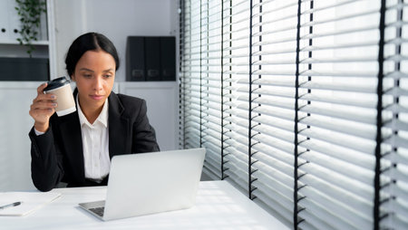 Competent female employee sits at her desk with a cup of coffee. Modern employee working with a...