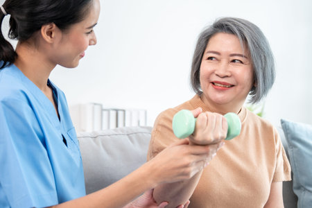 Caregiver helping contented senior woman exercise with dumbbell at home. Rehab for elder, senior...