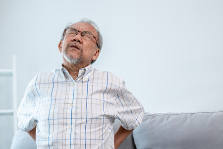 An agonizing senior man in need of assistance while sitting on his sofa at home, suffering from back pain. Senior care, nursing home for pensioners, deteriorating health of old age.の写真素材