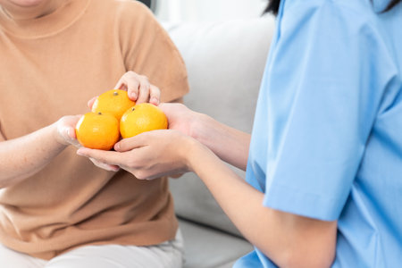 A young caregiver handing oranges to her contented senior patient at the living room. Senior care services, home visit by medical.の写真素材