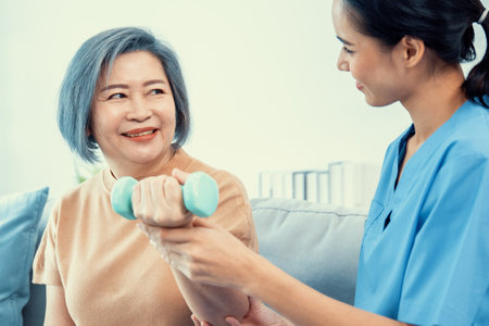 Caregiver helping contented senior woman exercise with dumbbell at home. Rehab for elder, senior medical care.の写真素材
