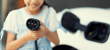 Focus electric charger plug from home charging station for EV engine power by renewable energy with blur background of girl pointing charger device at camera. Concept of progressive young generation.の写真素材