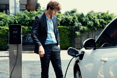 Progressive businessman wearing black suit with electric car recharging at public parking car charging station at modern city residential area. Eco friendly rechargeable car powered by clean energy.の写真素材