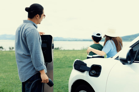 Concept of progressive happy family enjoying their time at green field and lake with electric vehicle. Electric vehicle driven by clean renewable from eco-friendly power sauce.の写真素材