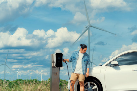 Progressive man with his electric car, EV car recharging energy from charging station on green field with wind turbine as concept of future sustainable energy. Electric vehicle with energy generator.の写真素材