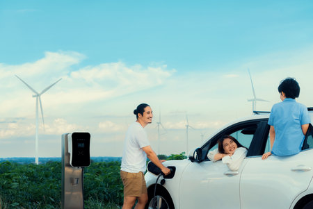 Concept of progressive happy family enjoying their time at wind farm with electric vehicle. Electric vehicle driven by clean renewable energy from wind turbine generator for charging station.の写真素材