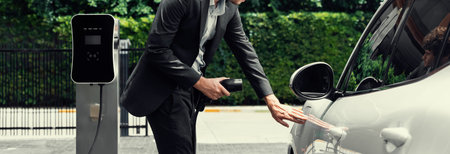 Progressive businessman with electric car recharging at public charging station at modern city...