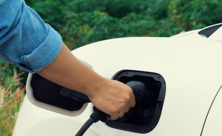 Progressive natural scenic where hand insert charging plug to electric vehicle from charging station with natural background. Future energy concept of EV car powered by sustainable electric energy.の写真素材
