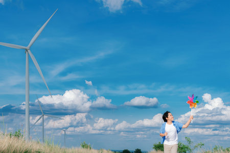 Progressive young asian boy playing with wind pinwheel toy in the wind turbine farm, green field over the hill. Green energy from renewable electric wind generator. Windmill in the countryside conceptの写真素材