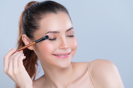 Closeup beautiful girl with flawless applying glamorous eye shadow makeup with eyeliner brush. Cosmetic facial painting process on lovely young woman with perfect clean skin in isolated background.の写真素材