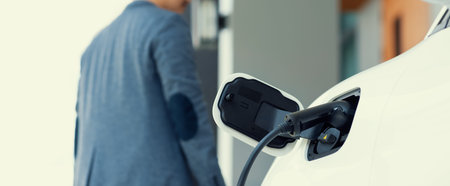 Focus electric car charging at home charging station with blurred progressive man walking in the...