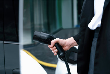 Focus EV charger plug and electric car at public charging station with blur progressive businesswoman holding charger and apartment condo building in background. Eco friendly electric vehicle concept.の写真素材