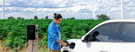 Progressive man with his electric car, EV car recharging energy from charging station on green field...