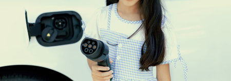 A playful girl holding and pointing an EV plug, a home charging station providing a sustainable power source for electric vehicles. Concept of progressive new generation with ecological awareness.の写真素材
