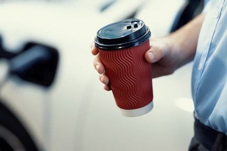 Progressive eco-friendly concept of parking EV car at public electric-powered charging station in city with blur background of businessman leaning on recharging-electric vehicle with coffee.の写真素材