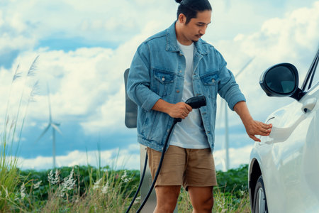 Progressive man with his electric car, EV car recharging energy from charging station on green field with wind turbine as concept of future sustainable energy. Electric vehicle with energy generator.の写真素材
