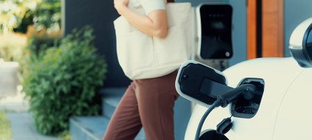 Focus image of electric vehicle recharging battery at home charging station with blurred woman walking in the background. Progressive concept of green energy technology applied in daily lifestyle.の写真素材