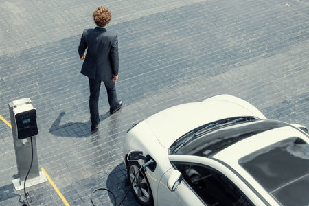 Aerial view of progressive businessman in black formal suit with his electric vehicle recharging battery at public car park charging station as vehicle powered by sustainable energy concept.の写真素材