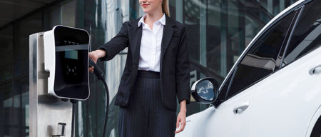 Progressive businesswoman wearing suit with electric car recharging at public parking car charging station at modern city center. Eco friendly rechargeable car powered by alternative clean energy.の写真素材